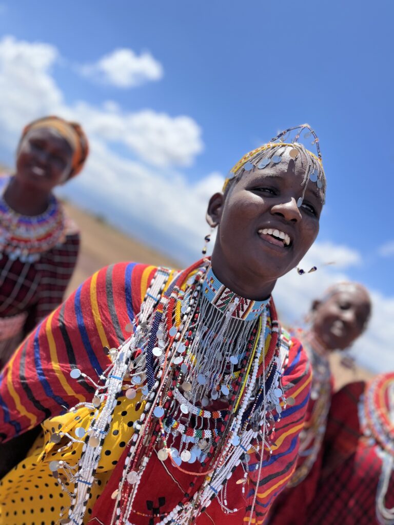 Portrait of a smiling Maasai woman adorned in traditional beaded jewelry and vibrant attire, set against a bright blue sky with others in similar dress in the background