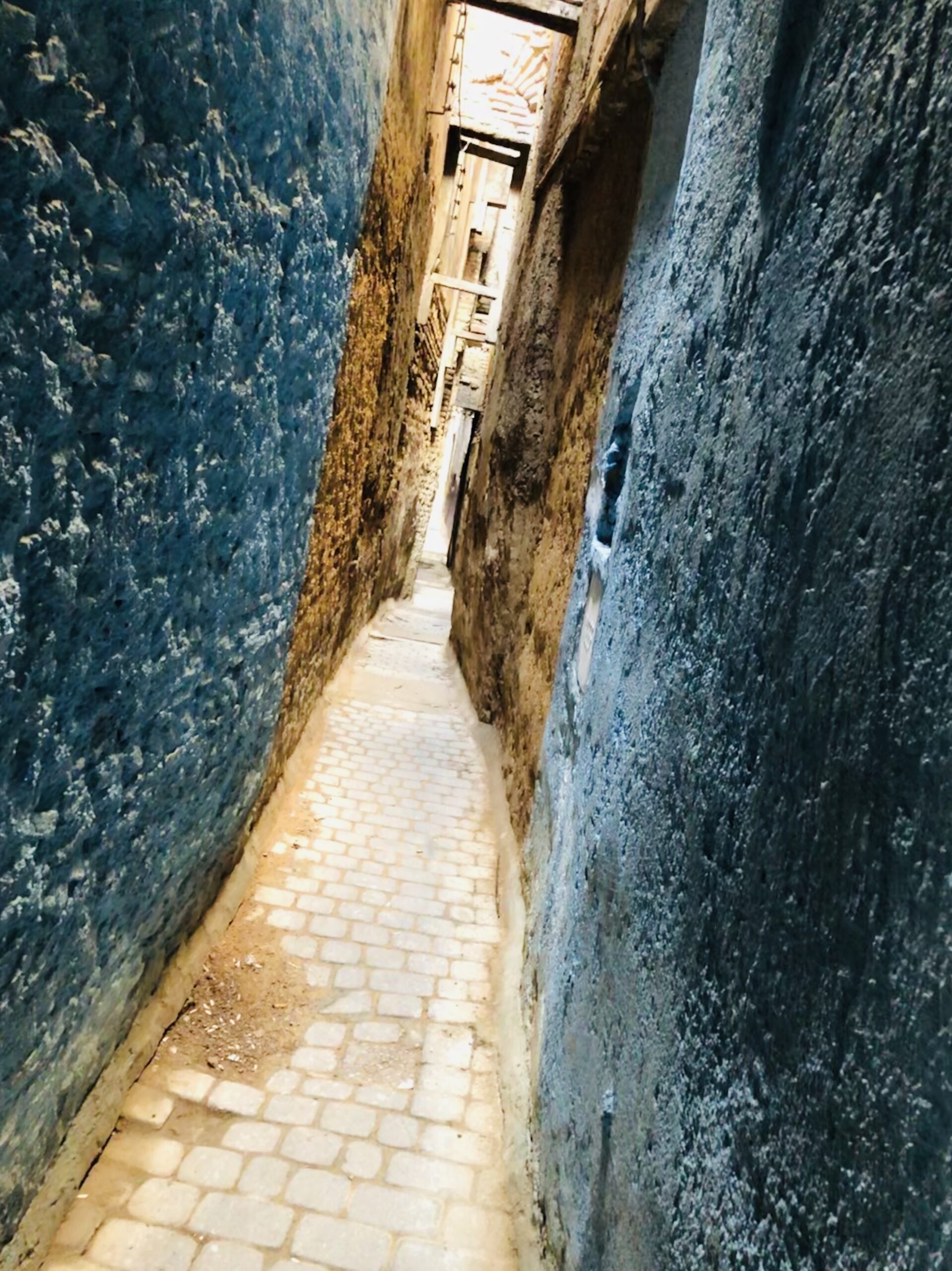 Extremely narrow cobblestone alleyway in the ancient medina of Fes, Morocco, with textured stone walls on either side and wooden beams overhead—one of the city’s famously tight passageways barely wide enough for a single person.
