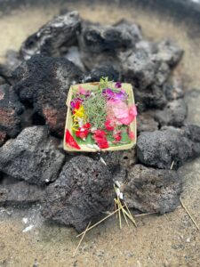 A traditional Balinese offering, known as a canang sari, sits atop a pile of black volcanic rocks. The small woven tray is filled with colorful flower petals, leaves, and bits of dried grass, used in daily rituals to honor the gods and maintain spiritual balance. Scattered incense sticks lie around the base.