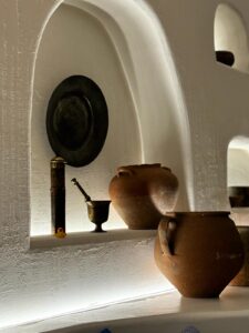 Rustic clay pots, a brass mortar and pestle, and a black metal plate are displayed in recessed white plaster shelves, softly illuminated by underlighting. A small yellow rubber duck adds a whimsical touch to the otherwise traditional decor.
