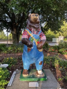 A painted bear statue wearing denim overalls and a red plaid shirt, standing upright in a garden bed surrounded by flowers and trees in New Bern, North Carolina.
