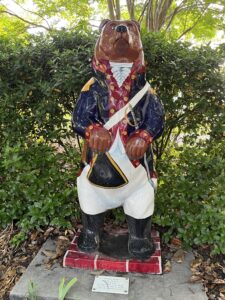 A bear statue dressed in a detailed Revolutionary War-era military uniform with red and gold accents, standing against a leafy green background in New Bern, North Carolina.