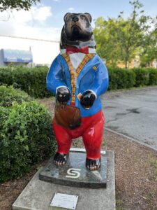 A bear statue styled as a well-dressed professional in a blue blazer, red bow tie, and red pants, standing on a sidewalk near shrubbery and a commercial building in New Bern, North Carolina.