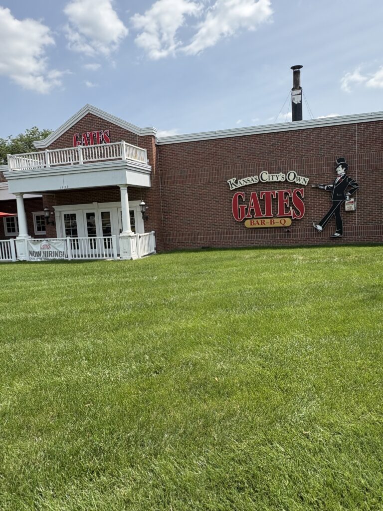 Exterior of Gates Bar-B-Q, a Kansas City barbecue staple, featuring a red brick building with white trim and a prominent sign that reads “Kansas City’s Own Gates Bar-B-Q.” A life-size illustration of a maître d’ in a tuxedo holding a tray decorates the wall. A well-manicured lawn stretches across the foreground under a sunny sky.
