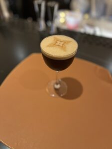 Close-up of a dark espresso martini on a tan leather placemat, featuring a foamy top with an intricate star-shaped design. The background is softly blurred, emphasizing the elegant cocktail presentation.