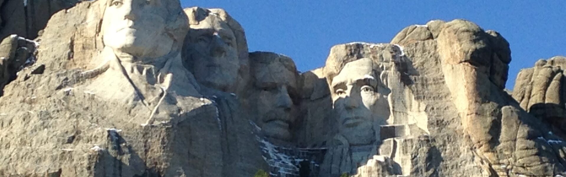Mount Rushmore National Memorial in South Dakota, featuring the carved faces of U.S. Presidents George Washington, Thomas Jefferson, Theodore Roosevelt, and Abraham Lincoln against a clear blue sky, with patches of snow and pine trees on the mountainside below.