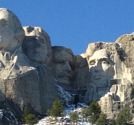 Mount Rushmore National Memorial in South Dakota, featuring the carved faces of U.S. Presidents George Washington, Thomas Jefferson, Theodore Roosevelt, and Abraham Lincoln against a clear blue sky, with patches of snow and pine trees on the mountainside below.