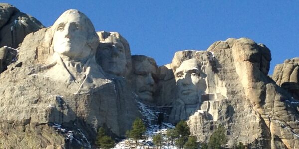 Mount Rushmore National Memorial in South Dakota, featuring the carved faces of U.S. Presidents George Washington, Thomas Jefferson, Theodore Roosevelt, and Abraham Lincoln against a clear blue sky, with patches of snow and pine trees on the mountainside below.