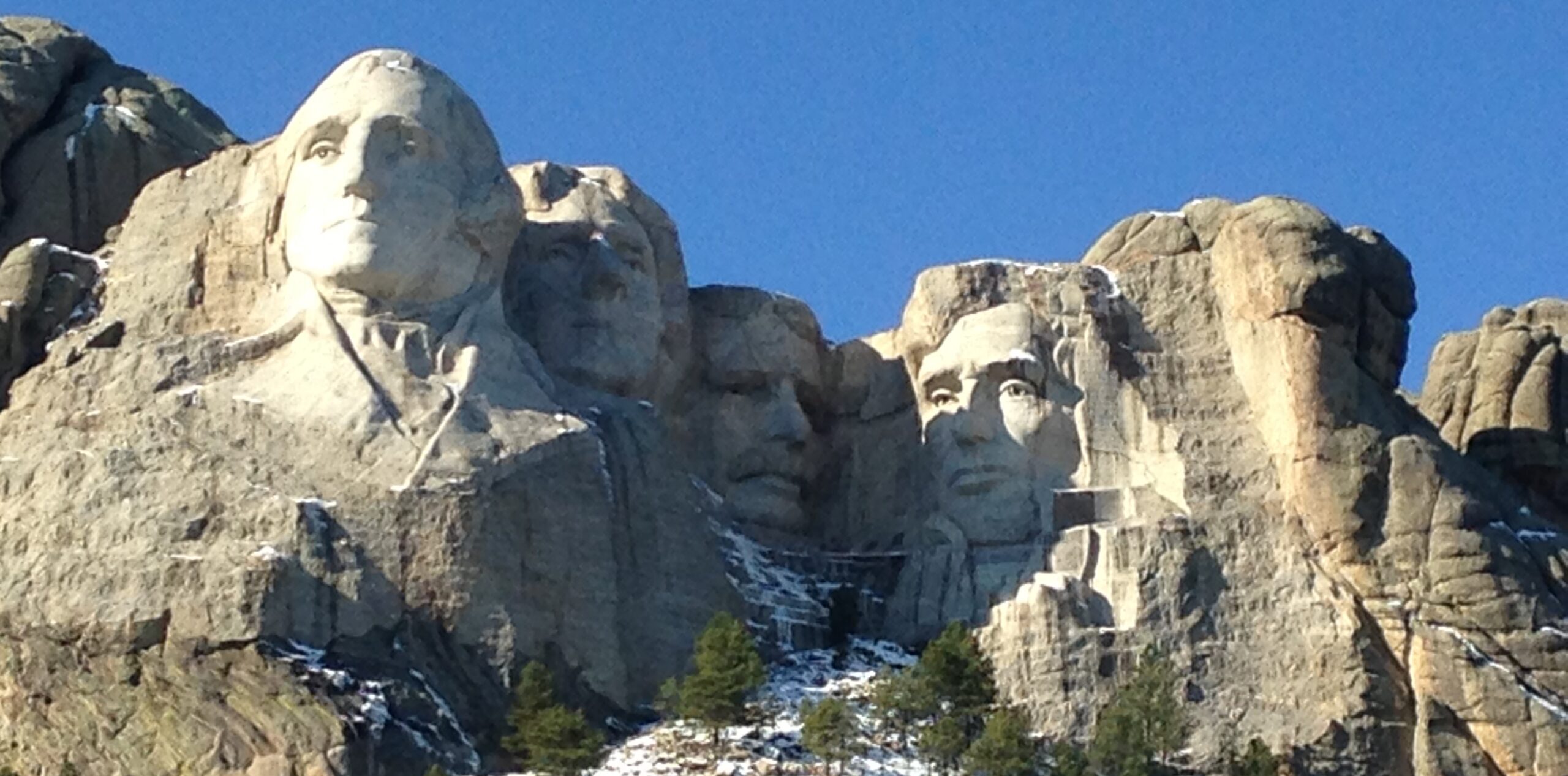 Mount Rushmore National Memorial in South Dakota, featuring the carved faces of U.S. Presidents George Washington, Thomas Jefferson, Theodore Roosevelt, and Abraham Lincoln against a clear blue sky, with patches of snow and pine trees on the mountainside below.