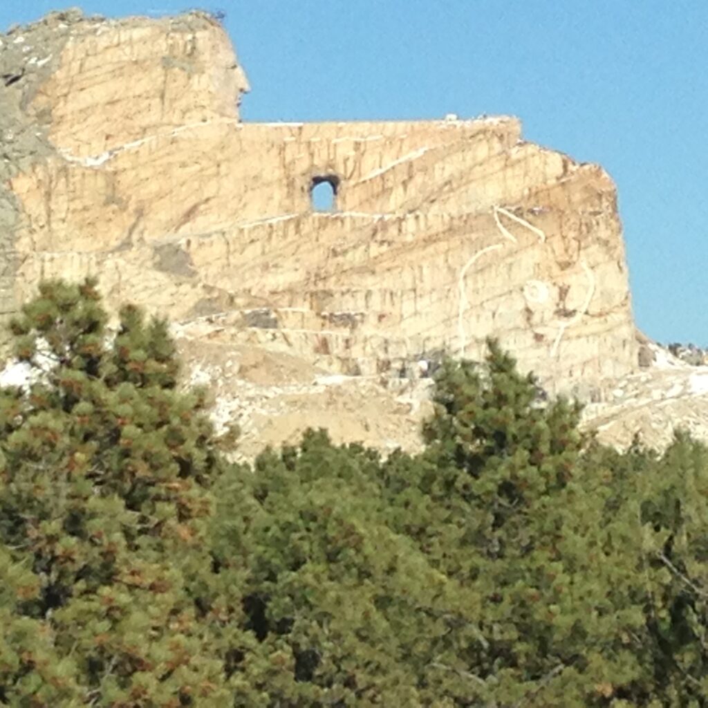 View of the Crazy Horse Memorial in South Dakota, an unfinished mountain carving depicting the Oglala Lakota warrior Crazy Horse pointing forward while riding a horse, set against a clear blue sky and partially surrounded by pine trees in the foreground.