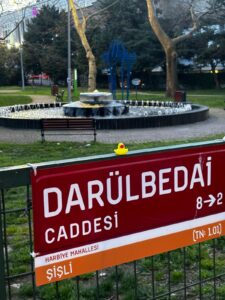 Street sign for Darülbedai Caddesi in the Harbiye neighborhood of Şişli, Istanbul, mounted on a green fence in front of a small park with a circular fountain and benches. A small yellow rubber duck playfully rests atop the sign.