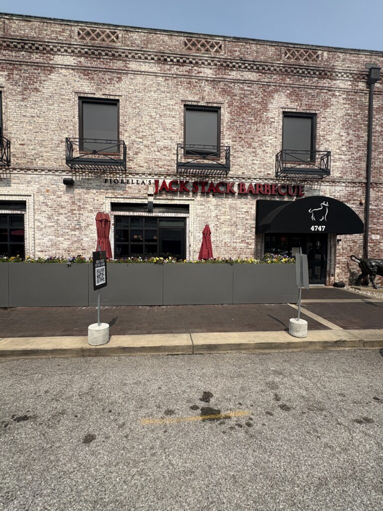 Front view of Fiorella’s Jack Stack Barbecue housed in a rustic brick building with black awnings and flower planters. Red umbrellas and an outdoor seating area line the sidewalk in front of the upscale barbecue restaurant, located at 4747.