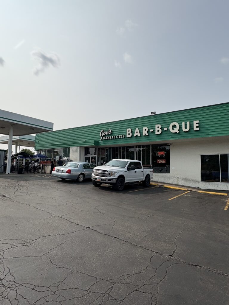 Exterior view of Joe's Kansas City Bar-B-Que located in a gas station with a green and white facade. A white pickup truck and a silver sedan are parked in front. A small group of people waits near the entrance under a gas station canopy on a sunny day.