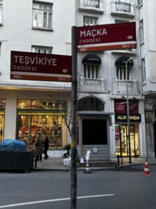 Street view of the intersection of Maçka Caddesi and Teşvikiye Caddesi in the Şişli district of Istanbul, Turkey. The scene features red street signs in Turkish, a shop window displaying colorful goods from MacKenzie-Childs, pedestrians, and historic European-style buildings in the background.