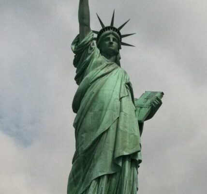 The Statue of Liberty on Liberty Island in New York Harbor, with its iconic green patina, torch raised high, and stone pedestal beneath a cloudy sky.