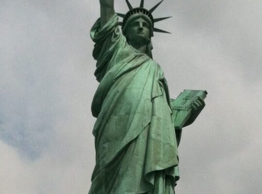 The Statue of Liberty on Liberty Island in New York Harbor, with its iconic green patina, torch raised high, and stone pedestal beneath a cloudy sky.
