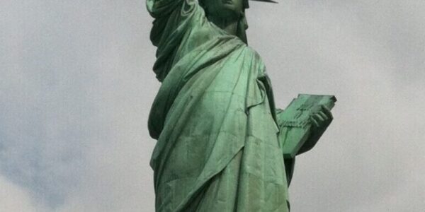 The Statue of Liberty on Liberty Island in New York Harbor, with its iconic green patina, torch raised high, and stone pedestal beneath a cloudy sky.