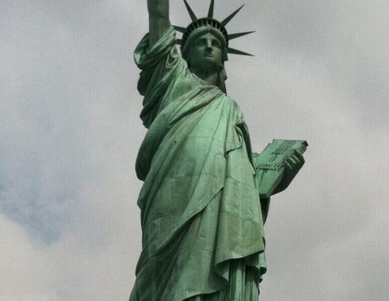 The Statue of Liberty on Liberty Island in New York Harbor, with its iconic green patina, torch raised high, and stone pedestal beneath a cloudy sky.