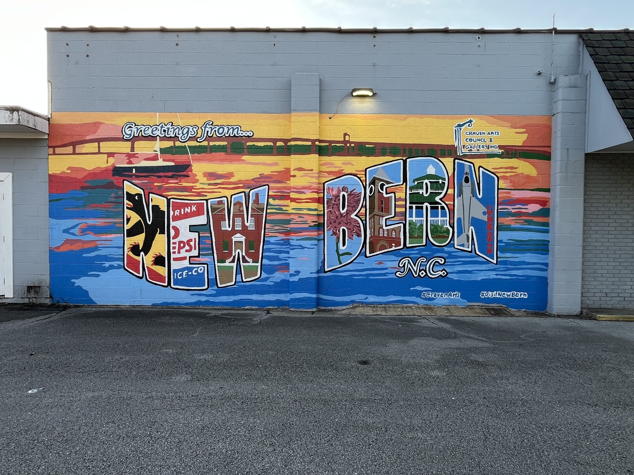 Colorful mural on a building wall reading “Greetings from New Bern, N.C.” Each letter is filled with local imagery including a Pepsi logo, waterfront scenes, and historic landmarks. The background features a vivid sunset over water with a sailboat and bridge. Tags include #CravenArts and #VisitNewBern.