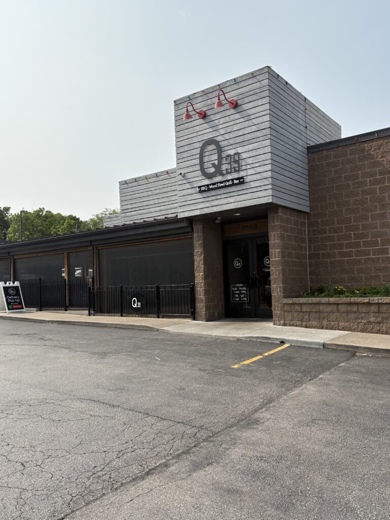 Exterior of Q39, a modern barbecue joint in Kansas City. The facade features a mix of gray siding and brown brick with bold black signage that reads “Q39 BBQ • Wood Fired Grill • Bar.” The entrance is marked by red overhead lights and a small patio area with a black metal fence.