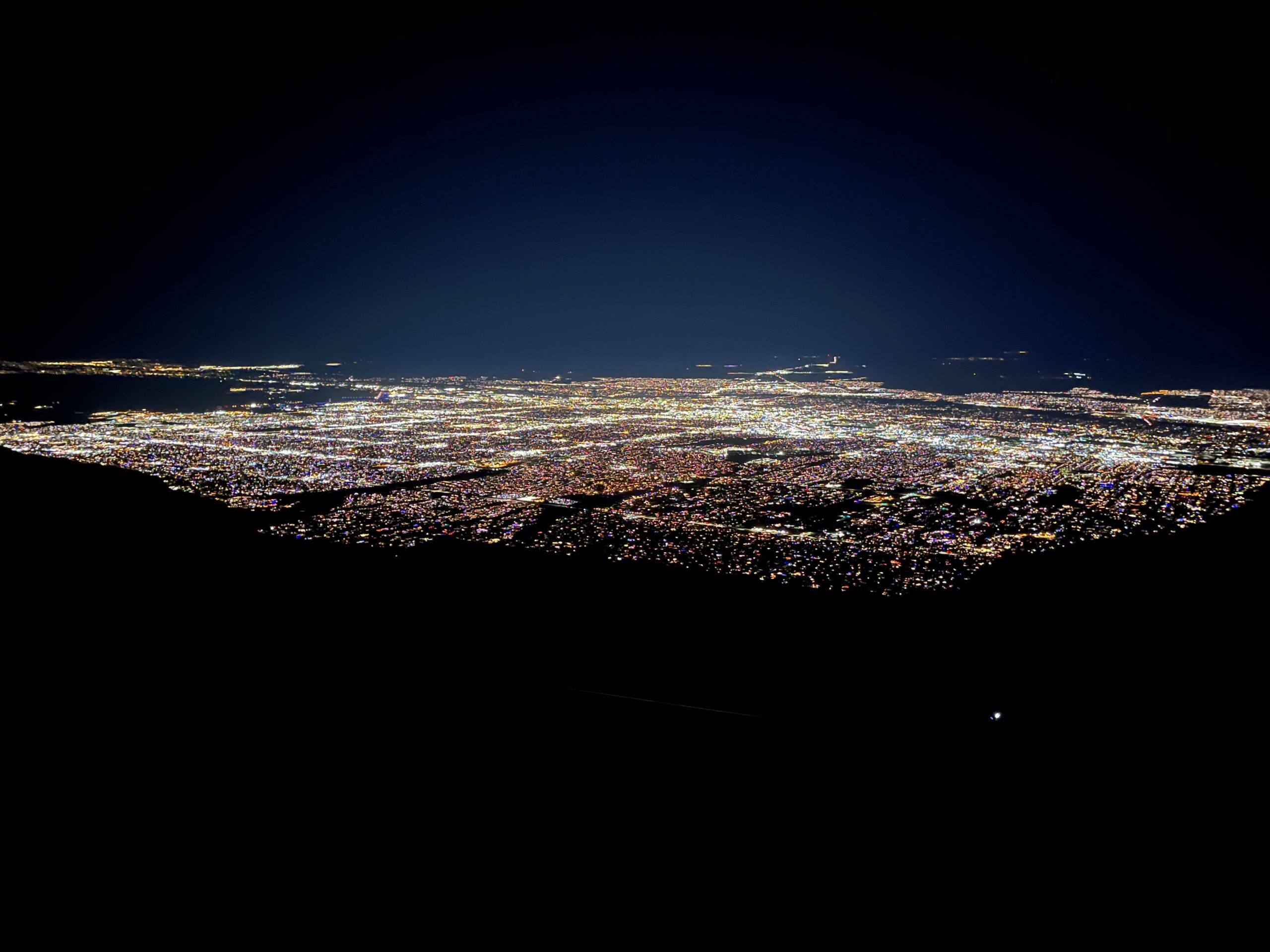 Nighttime view of Albuquerque, New Mexico, from the top of Sandia Peak. The city glows with a sea of colorful lights stretching across the valley, seen from a high, dark vantage point, highlighting the vast urban landscape against a clear night sky.