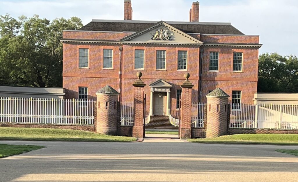 Front view of the historic Tryon Palace in New Bern, North Carolina, a red-brick Georgian-style mansion with symmetrical windows and a grand entrance flanked by round brick gateposts. A wide gravel path leads up to the stately building, framed by white fencing and green lawns.
