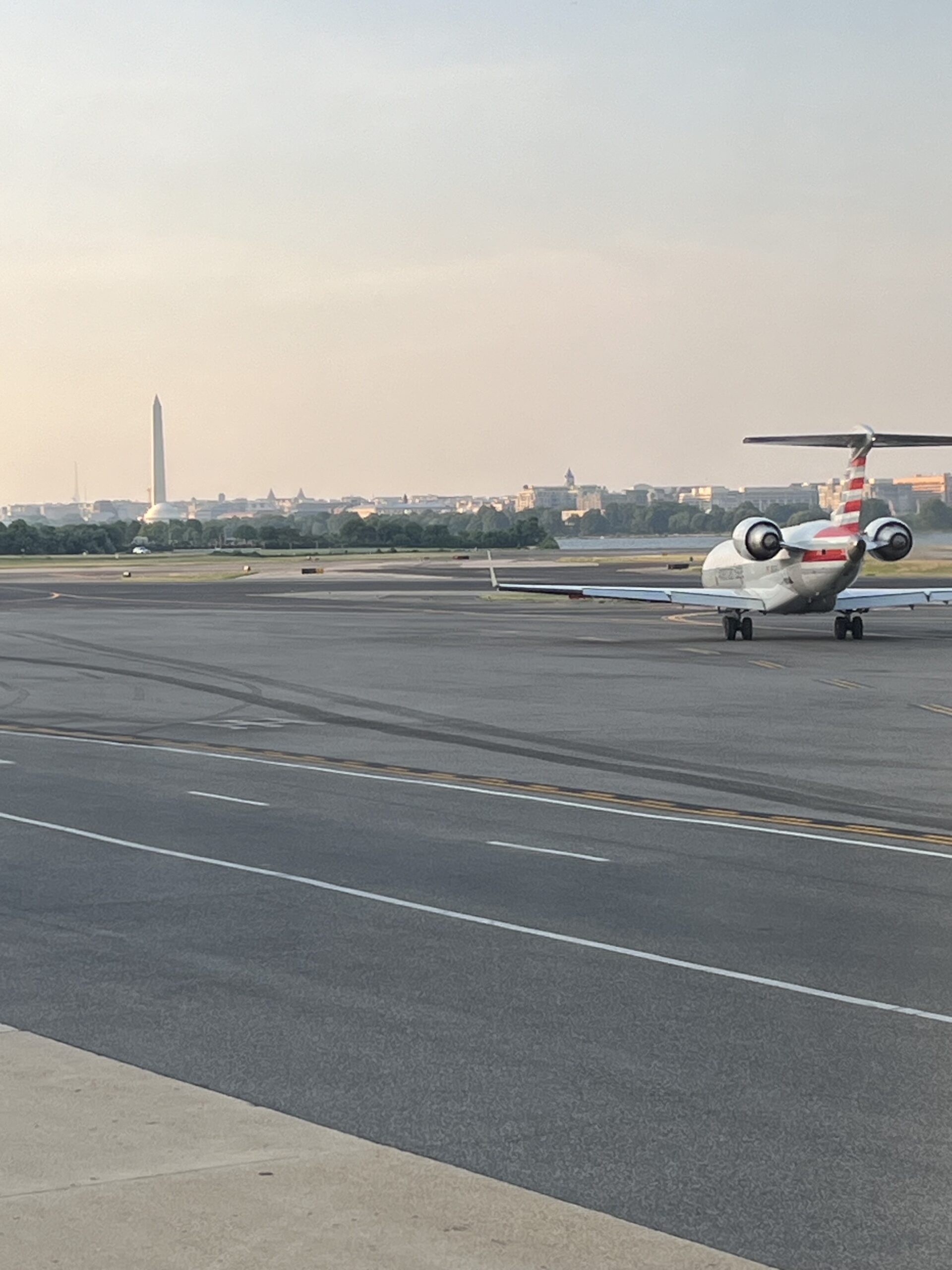 An American Airlines regional jet taxiing on the runway at Washington National Airport (DCA), with the Washington Monument and city skyline visible in the distance under a hazy sky.