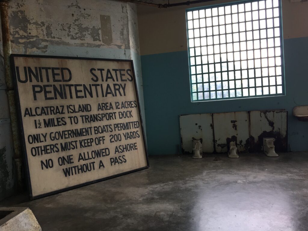 A large, weathered sign inside Alcatraz reading "United States Penitentiary," displayed beside rusted sinks and barred windows, capturing the stark, decaying interior of the former prison.