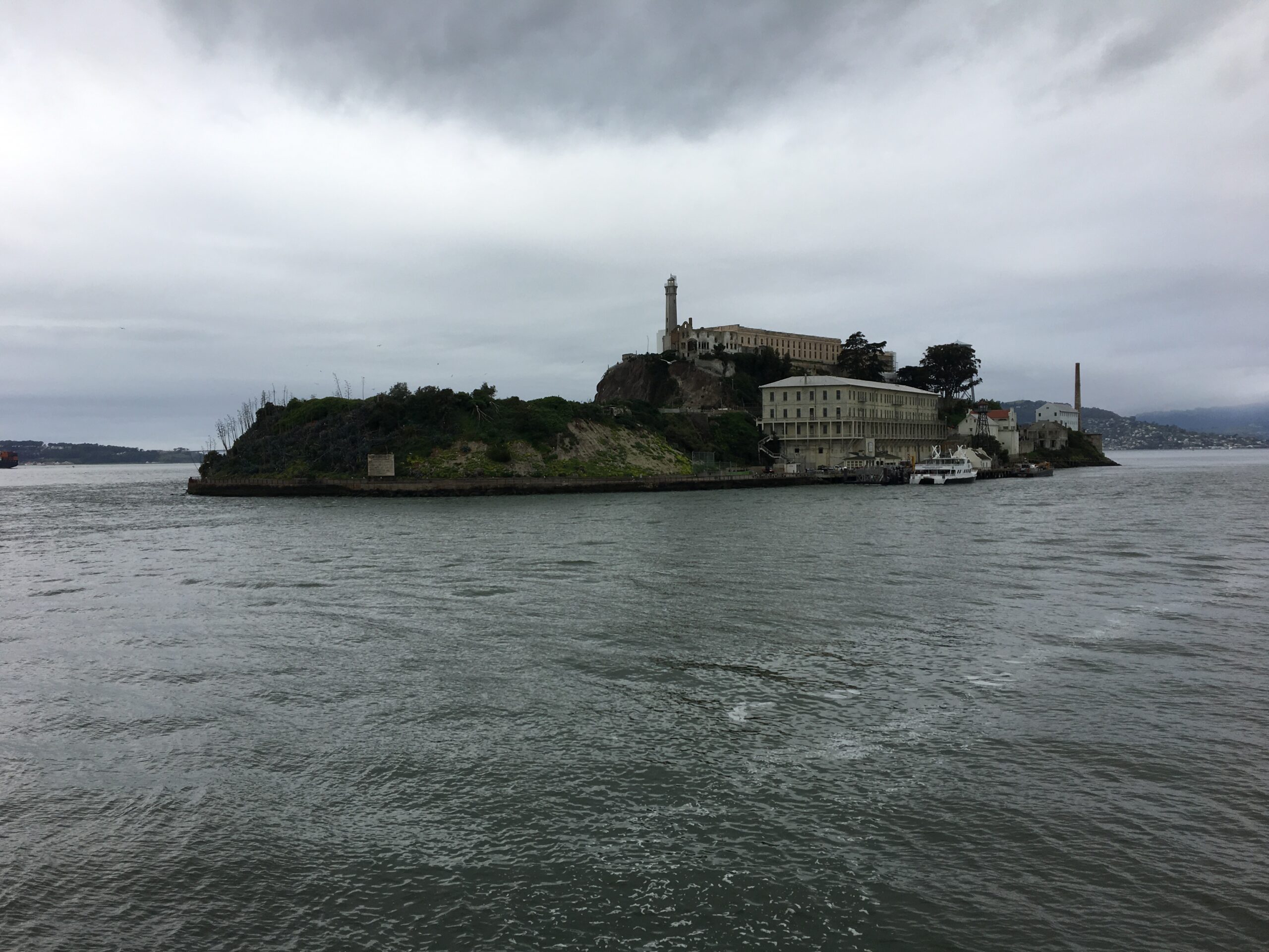 Distant view of Alcatraz Island under a cloudy sky, showing the former prison buildings and lighthouse surrounded by cold, choppy waters of the San Francisco Bay.