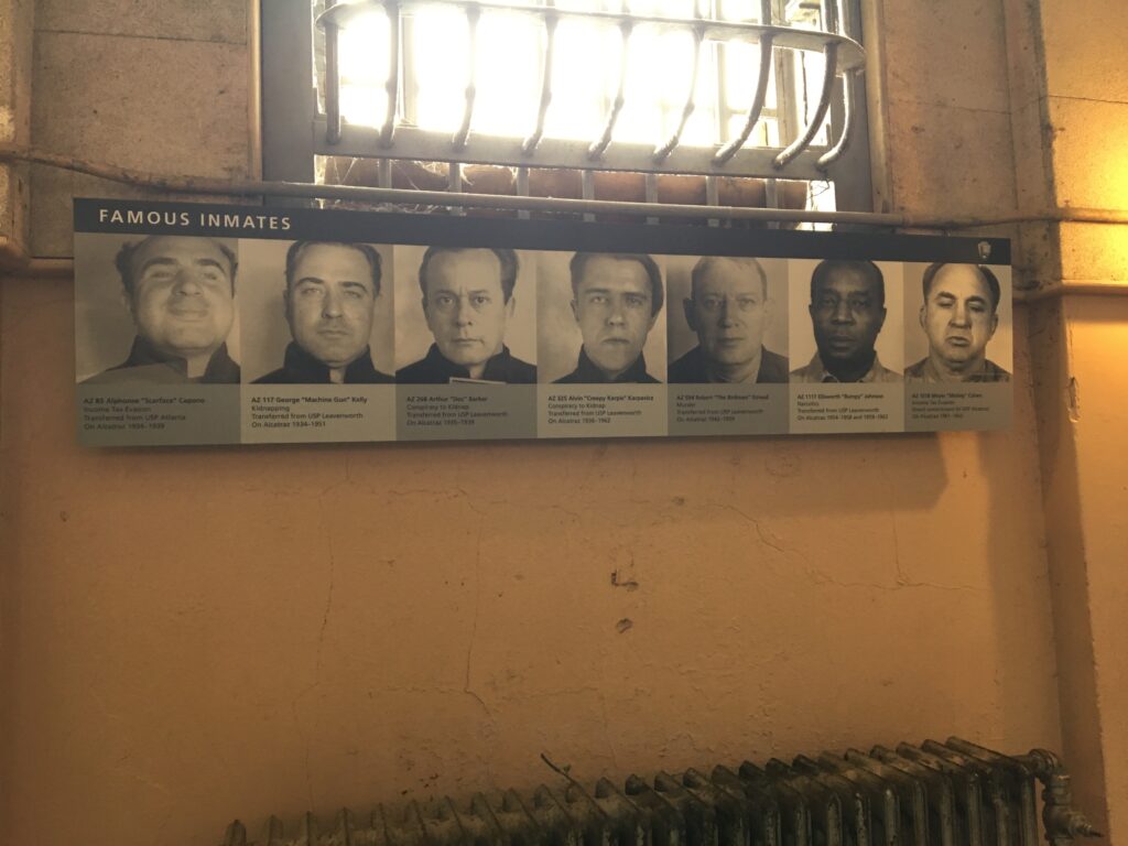 Interior display at Alcatraz featuring black-and-white mugshots of infamous inmates, including Al Capone, George “Machine Gun” Kelly, and Ellsworth “Bumpy” Johnson, mounted beneath a barred window on a worn prison wall above a rusted radiator—highlighting the notorious history of the island penitentiary.