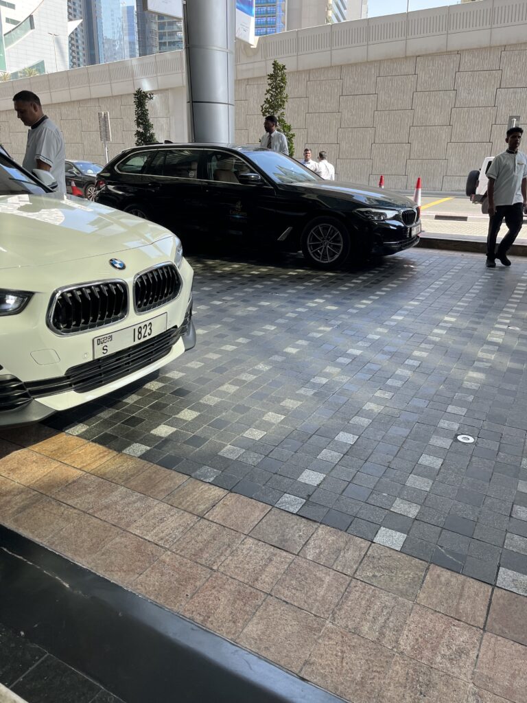 Valet area in front of a luxury hotel in Dubai, showing a white BMW SUV and a black BMW sedan with an "Emirates Chauffeur Drive" logo. Several uniformed staff members stand nearby, with high-rise buildings and a stone wall in the background.