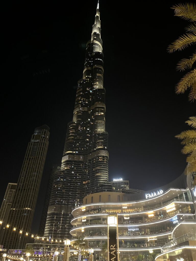 The Burj Khalifa illuminated against the night sky in downtown Dubai, with the glowing EMAAR mall and palm trees in the foreground, surrounded by twinkling string lights.
