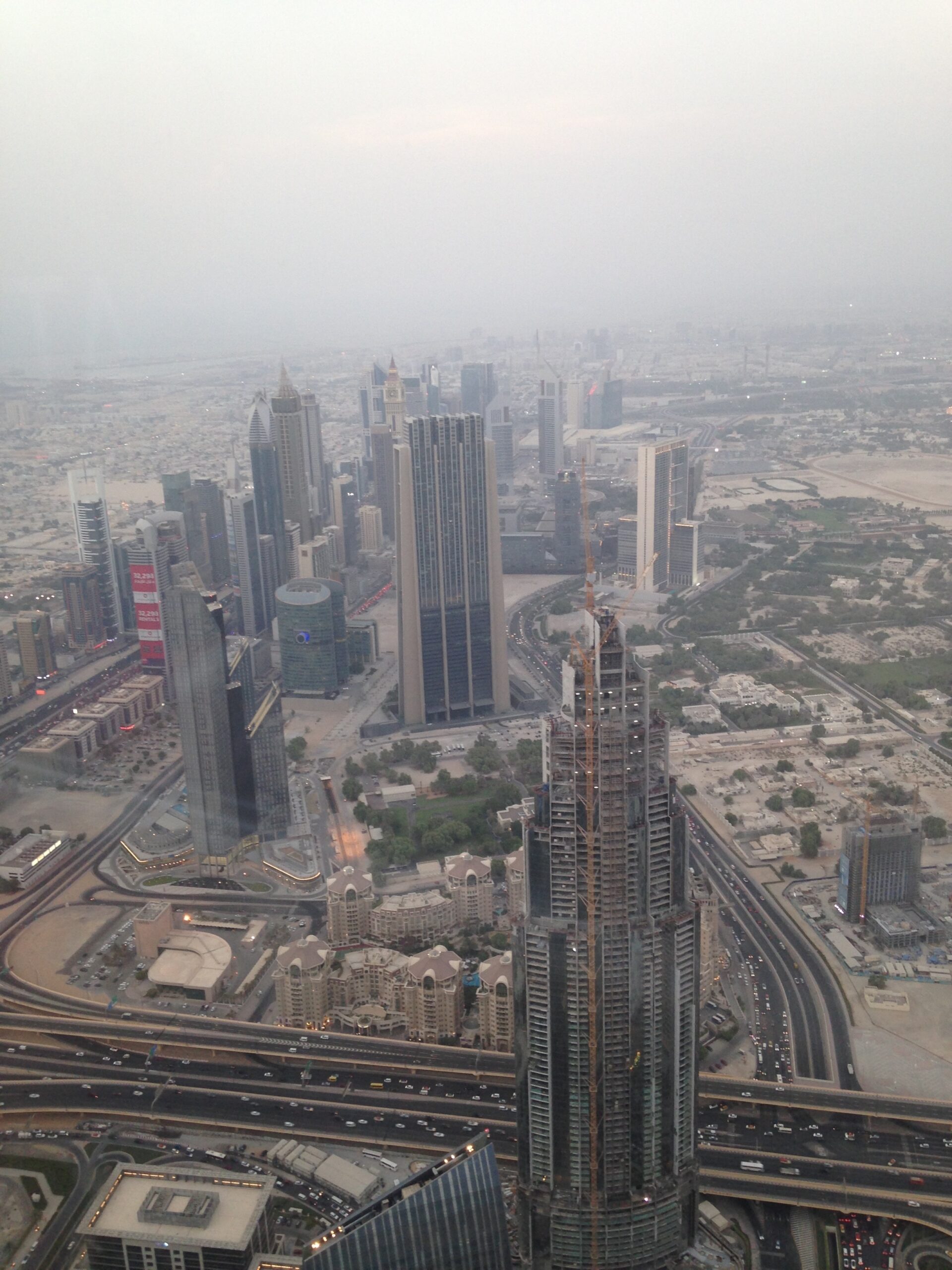 A hazy aerial view of Dubai's skyscrapers and highways, taken from a high vantage point, showcasing modern architecture, dense urban planning, and desert landscape in the background.