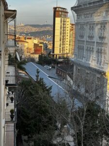 View from a hotel balcony in Istanbul overlooking a tree-lined courtyard and residential buildings, with a modern high-rise and the distant skyline of the city, including the Bosphorus and a large Turkish flag in the background.