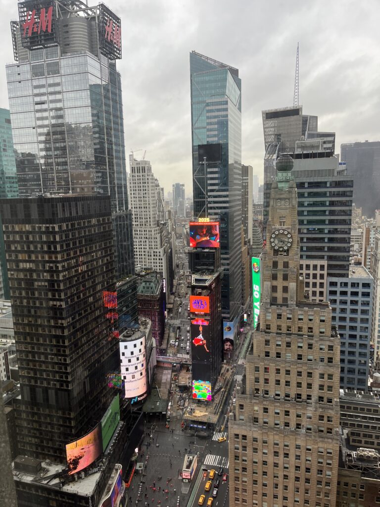 A high-rise view of Times Square in New York City from the Marriott Marquis hotel, showcasing electronic billboards, taxis, and surrounding skyscrapers on a cloudy day.
