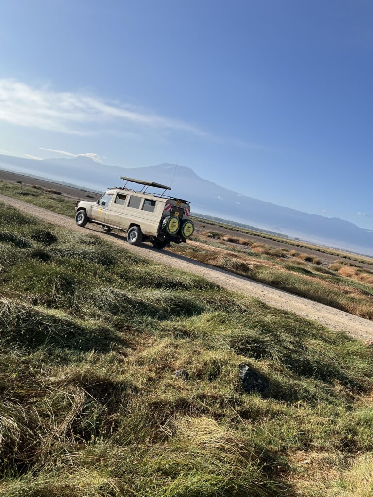 A beige safari jeep with a pop-up roof is parked on a dirt road in Amboseli National Park, Kenya. Tall grasses surround the vehicle, and the majestic silhouette of Mount Kilimanjaro rises in the background under a clear blue sky.