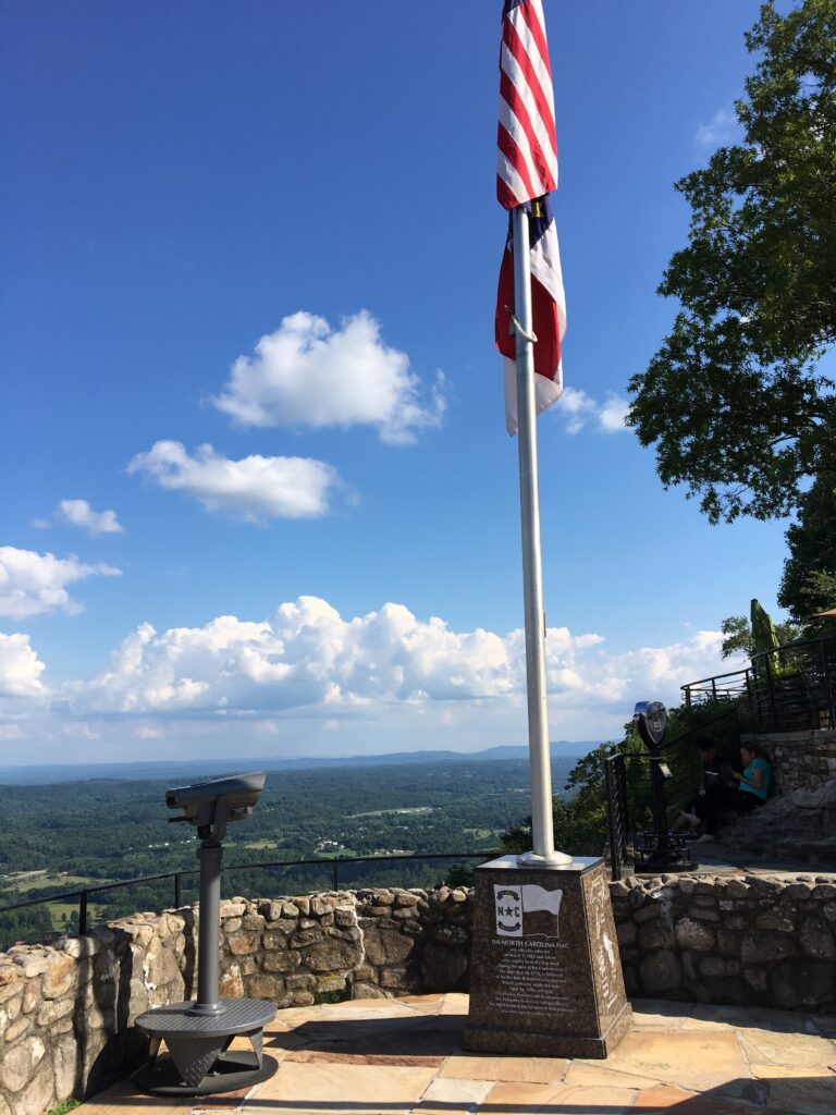 Scenic overlook at Lookout Mountain featuring a tall flagpole flying the U.S. and North Carolina flags, with a panoramic view of rolling hills and a stone marker honoring the state, all beneath a bright blue sky with fluffy clouds.
