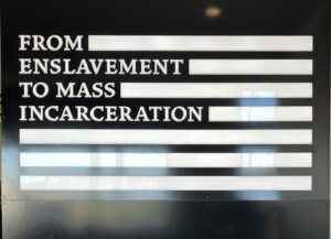 A wall display at the Legacy Museum in Montgomery, Alabama, reads “From Enslavement to Mass Incarceration” in bold white letters against a black background, with bars resembling a stylized American flag.