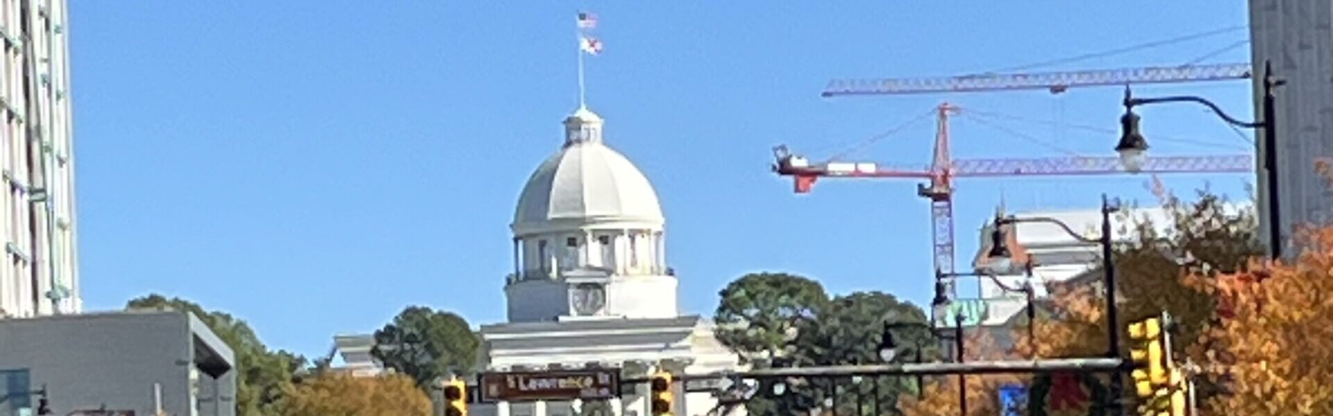 A clear daytime view of Dexter Avenue in Montgomery, Alabama, leading up to the Alabama State Capitol building with its prominent white dome and American flag on top. The street is lined with cars and autumn-colored trees, while red construction cranes are visible in the background.
