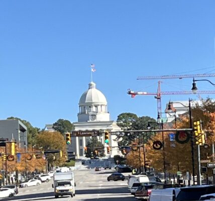 A clear daytime view of Dexter Avenue in Montgomery, Alabama, leading up to the Alabama State Capitol building with its prominent white dome and American flag on top. The street is lined with cars and autumn-colored trees, while red construction cranes are visible in the background.