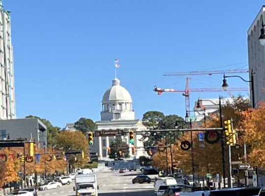 A clear daytime view of Dexter Avenue in Montgomery, Alabama, leading up to the Alabama State Capitol building with its prominent white dome and American flag on top. The street is lined with cars and autumn-colored trees, while red construction cranes are visible in the background.