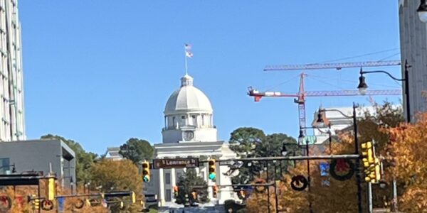 A clear daytime view of Dexter Avenue in Montgomery, Alabama, leading up to the Alabama State Capitol building with its prominent white dome and American flag on top. The street is lined with cars and autumn-colored trees, while red construction cranes are visible in the background.