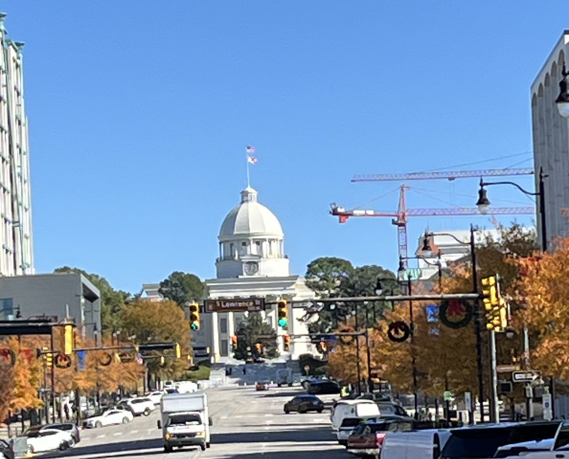 A clear daytime view of Dexter Avenue in Montgomery, Alabama, leading up to the Alabama State Capitol building with its prominent white dome and American flag on top. The street is lined with cars and autumn-colored trees, while red construction cranes are visible in the background.