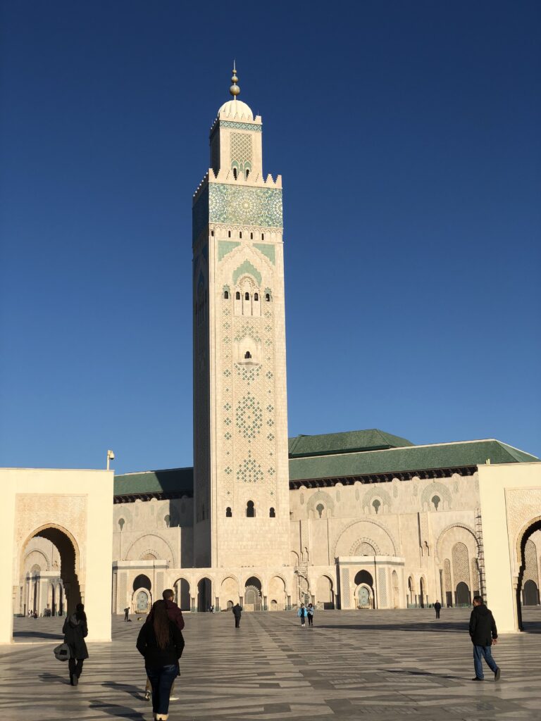 The towering minaret of the Hassan II Mosque in Casablanca, Morocco, rises against a clear blue sky as visitors stroll across the vast marble courtyard of one of the world’s largest and most stunning mosques.