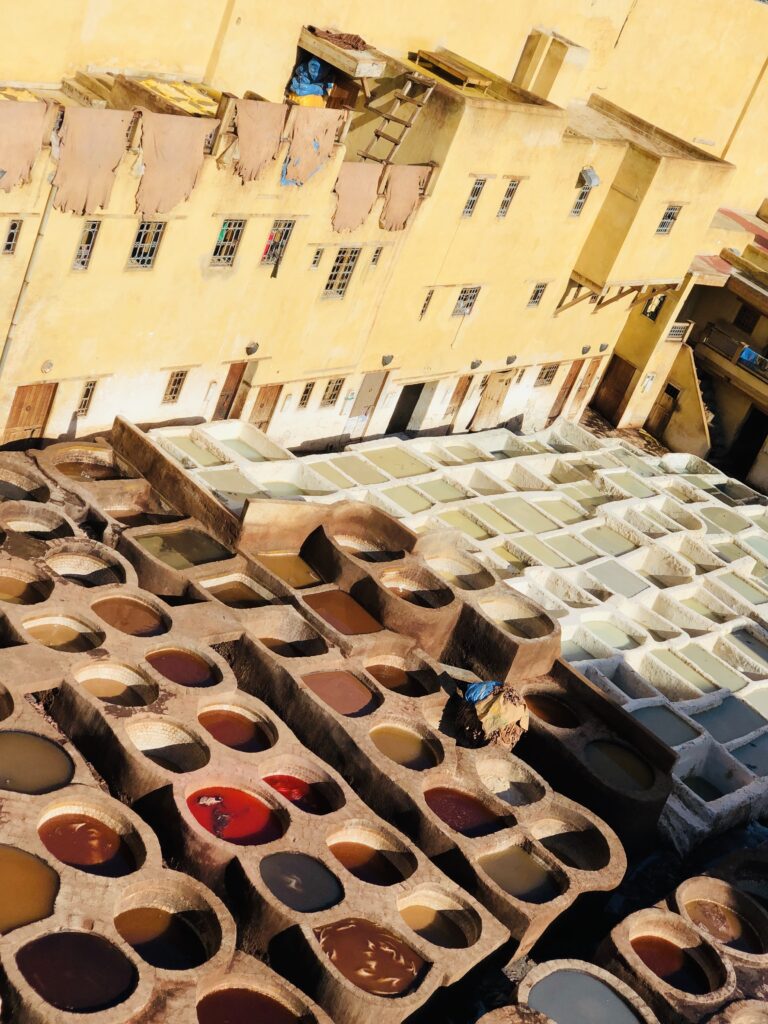 Vibrant view of the traditional leather tanning pits in Fes, Morocco, showing rows of stone vats filled with colorful natural dyes and soaking agents, with animal hides drying on the rooftop of a yellow building in the background—a centuries-old artisan process still in use today.