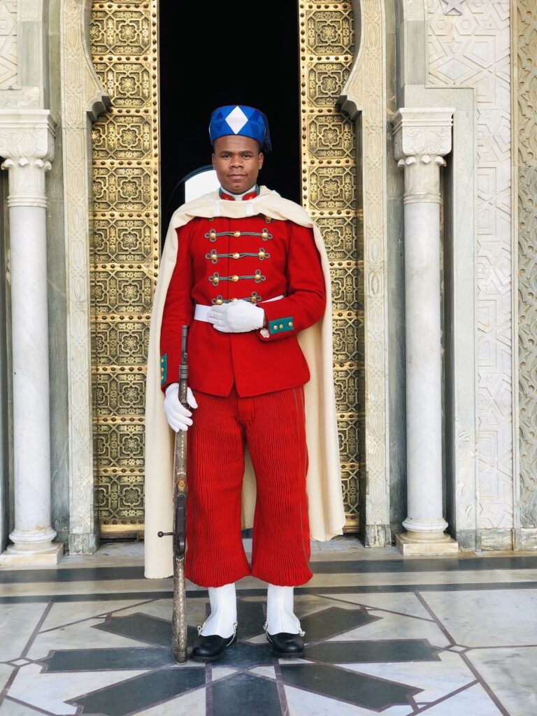 Moroccan royal guard in full ceremonial uniform—red jacket with gold buttons, pleated trousers, white gloves, and a blue hat—standing in front of an ornate golden door at the Mausoleum of Mohammed V in Rabat, Morocco.