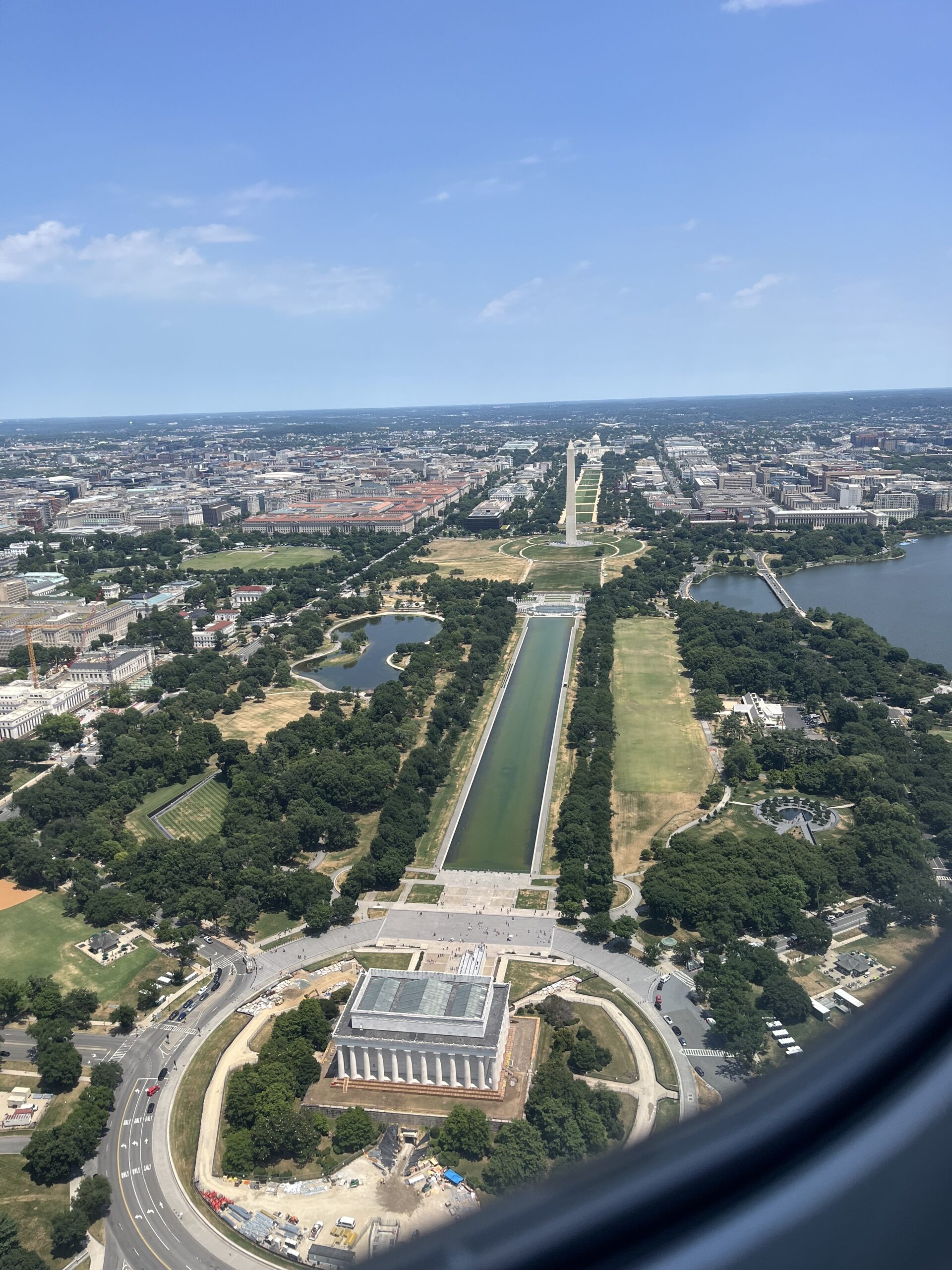 Aerial view of the National Mall in Washington, D.C., prominently featuring the Lincoln Memorial in the foreground, the Reflecting Pool, and the Washington Monument aligned in the center, with the U.S. Capitol visible in the distance under a clear blue sky.
