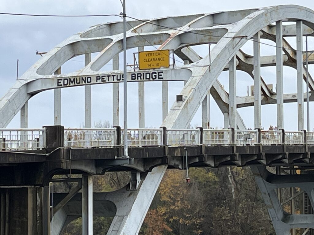 A close-up view of the historic Edmund Pettus Bridge in Selma, Alabama. The arched steel structure, weathered with age, spans the Alabama River and features a yellow sign indicating vertical clearance. This site is famously known for the 1965 Selma to Montgomery marches during the Civil Rights Movement.