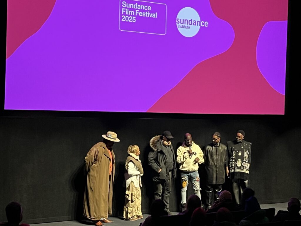 A panel of six speakers stands onstage in front of a large screen displaying the Sundance Film Festival 2025 and Sundance Institute logos. The group, dressed in distinctive and stylish winter attire, engages with the audience during a post-screening discussion. The theater is dimly lit, with audience silhouettes visible in the foreground.