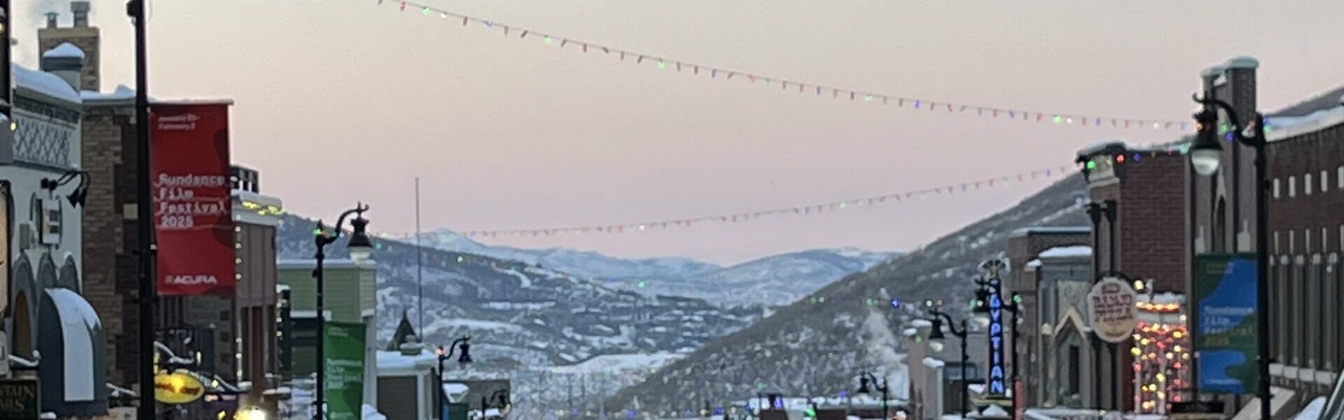 Main Street in Park City, Utah, is bustling with pedestrians during the 2025 Sundance Film Festival. Snow lines the sidewalks, festive string lights hang above the street, and colorful banners and storefronts glow under the early evening sky with mountains in the distance.